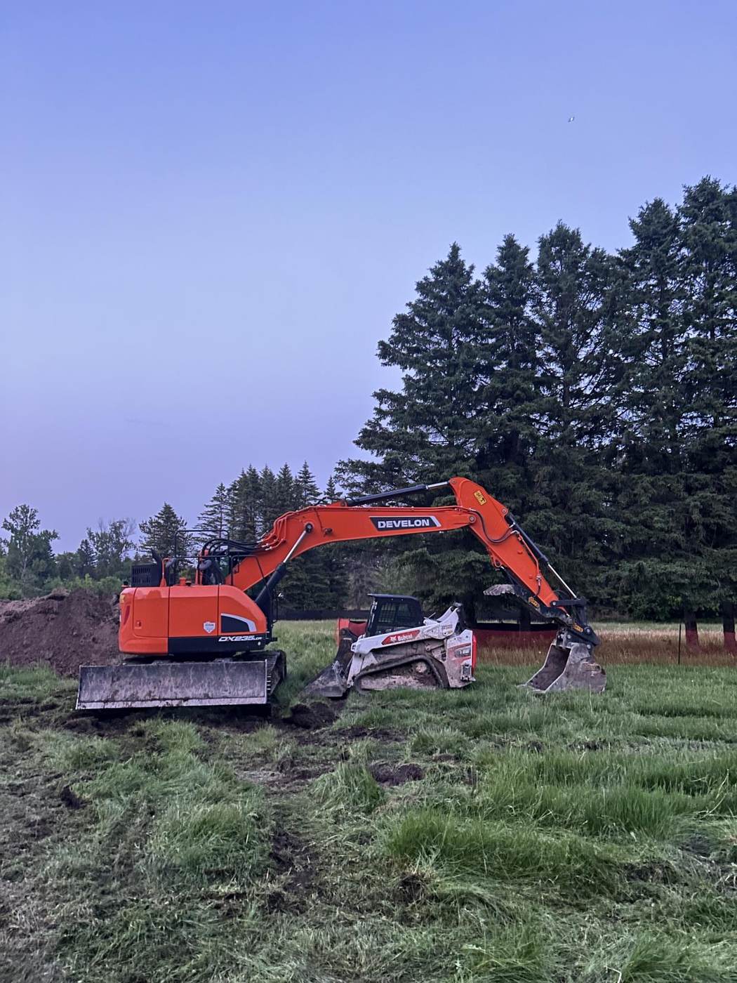 Orange Develon DX235 excavator beside a Bobcat track loader at dusk
